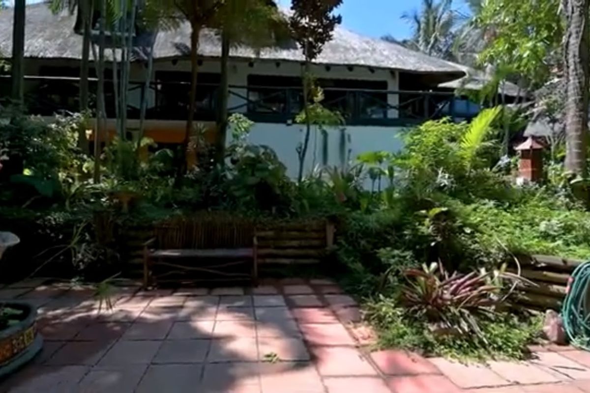 A rear view of the property featuring a thatched roof, a balcony, and a paved patio area surrounded by lush garden landscaping.