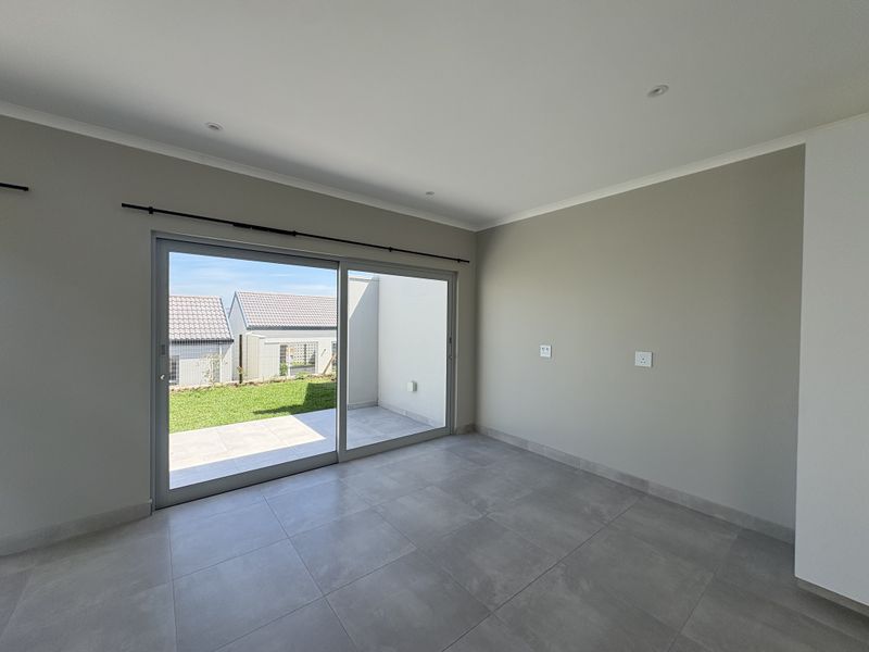 A bright, empty living room featuring large sliding glass doors that open onto a private patio and garden area, finished with modern floor tiles.