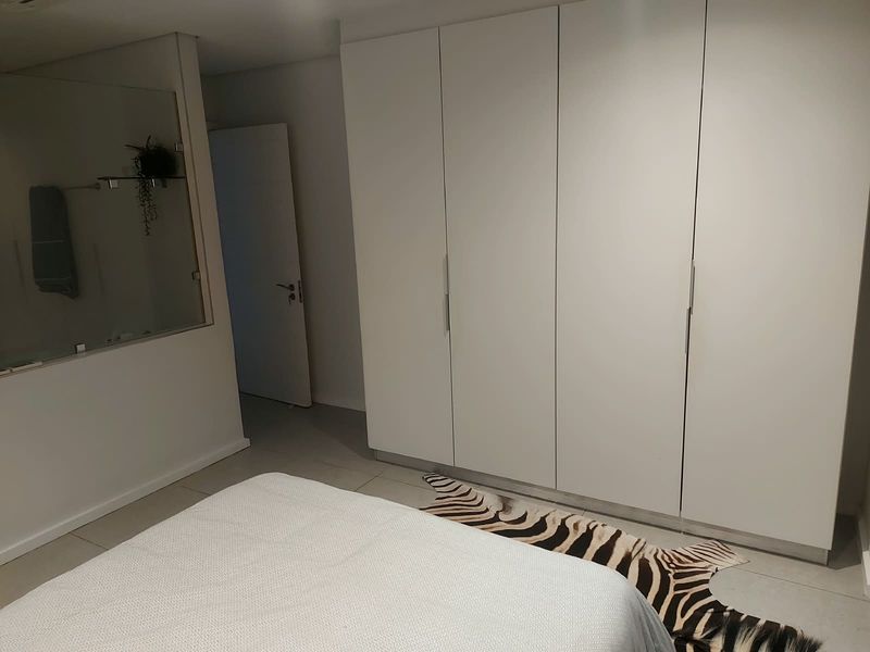 A modern bedroom featuring sleek white built-in wardrobes, neutral tiled flooring, and a unique internal glass partition wall.