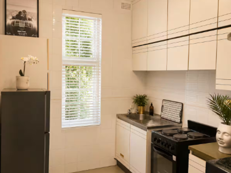 A compact kitchen featuring white cabinetry, a stainless steel sink, a freestanding stove, and tiled walls, illuminated by a modern ceiling light fixture.