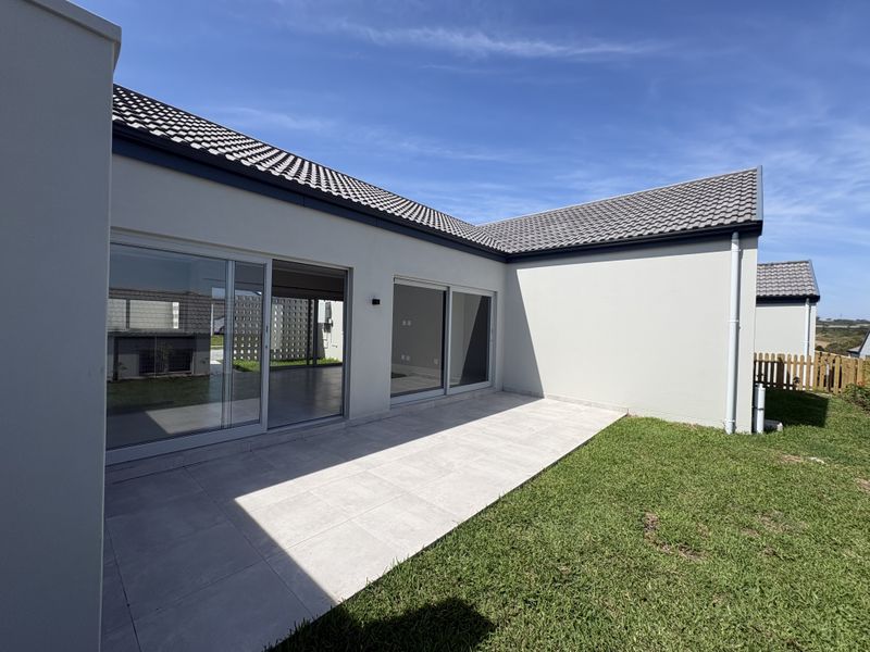A modern rear exterior view featuring a tiled patio, large sliding glass doors, and a well-maintained lawn under a clear blue sky.