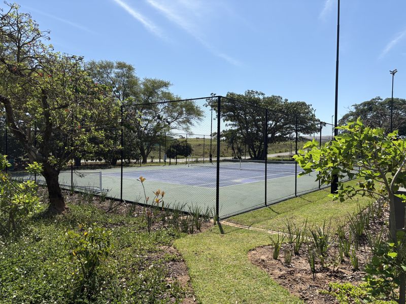 A well-maintained outdoor tennis court enclosed by chain-link fencing, situated within a landscaped estate setting.