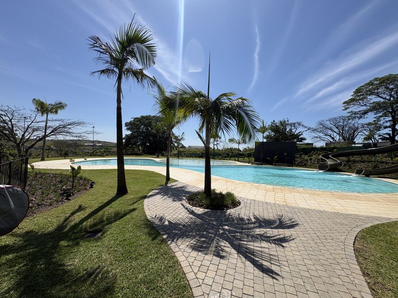 A well-maintained outdoor pool area featuring a paved deck, landscaped garden, and a built-in water slide, perfect for recreational use.