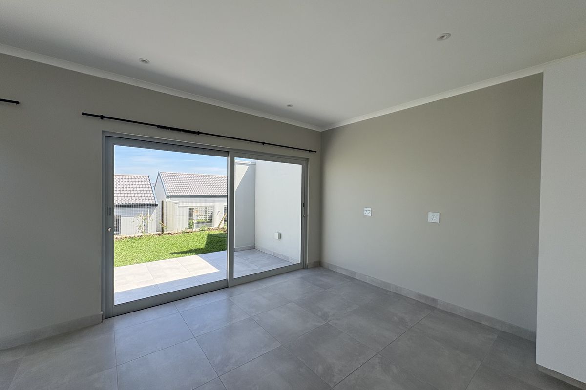 A bright, empty living room featuring large sliding glass doors that open onto a private patio and garden area, finished with modern floor tiles.