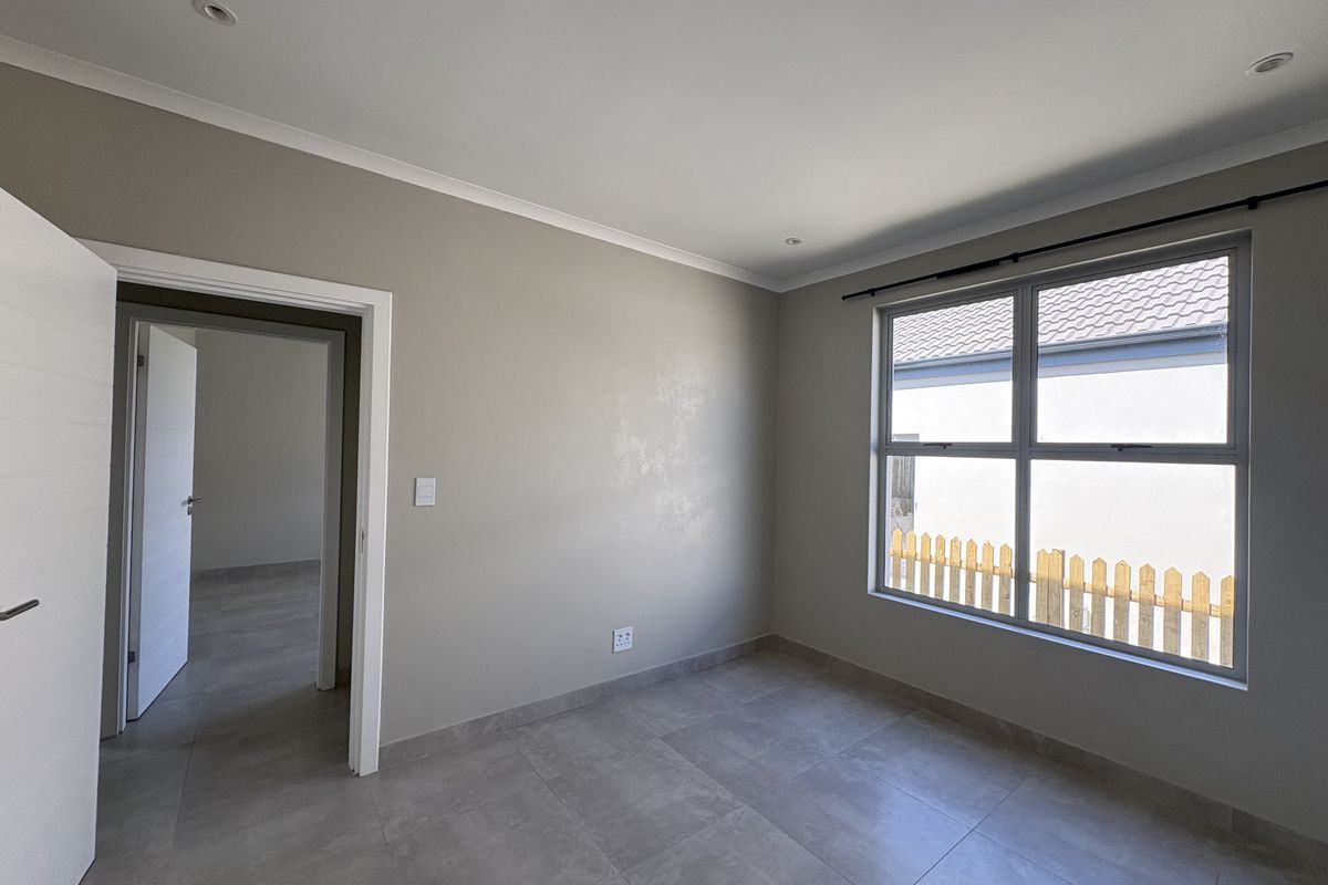 A bright, empty bedroom featuring neutral grey tiled flooring, recessed downlighting, and a large window with views of the exterior fence.