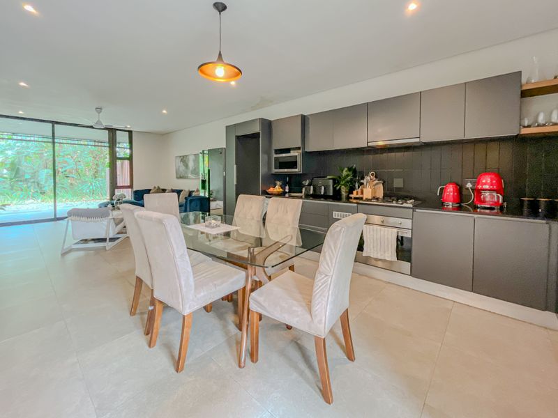 Modern open-plan kitchen and dining area featuring sleek dark cabinetry, integrated appliances, tiled flooring, and large sliding doors leading to an outdoor space.