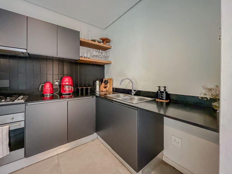 Modern kitchen featuring sleek dark cabinetry, stone countertops, integrated stainless steel sink, built-in oven, gas hob, and stylish floating timber shelving.