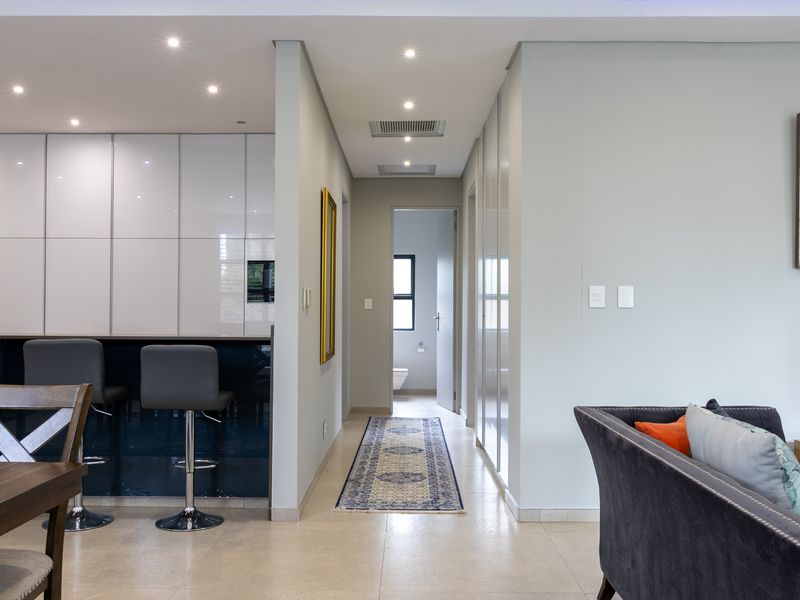 Modern open-plan living area featuring high-gloss kitchen cabinetry, a central island, recessed lighting, and seamless tiled flooring leading to a hallway.