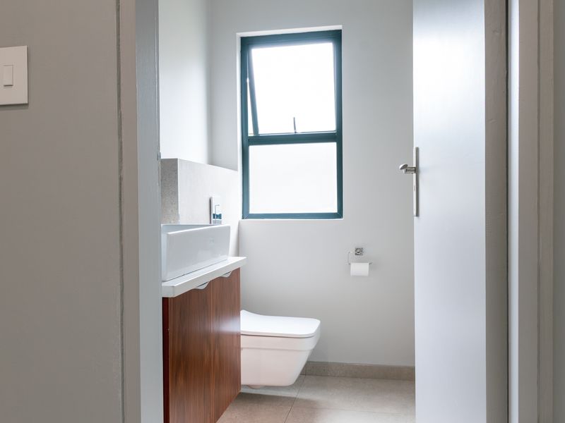 A clean, modern bathroom featuring a wall-mounted toilet, a wood-finish vanity with a vessel sink, and a functional casement window.
