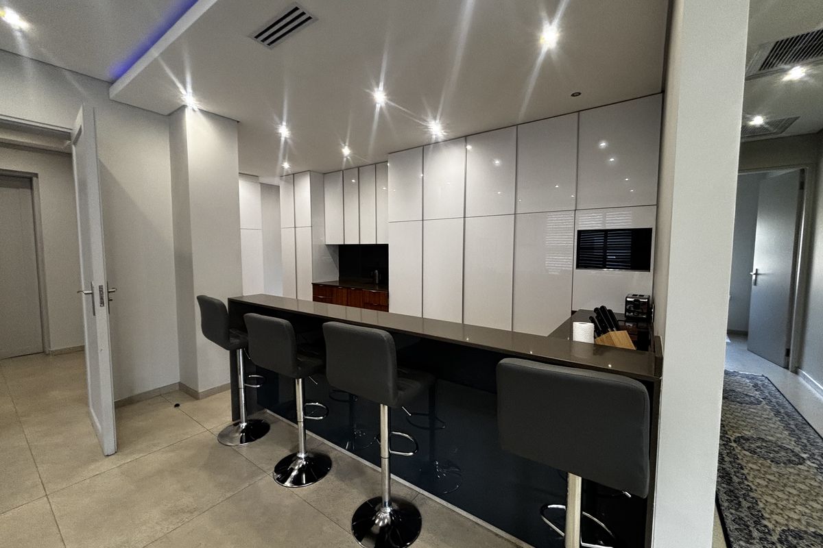 Modern kitchen featuring sleek white cabinetry, a dark stone breakfast bar, recessed ceiling lighting, and large format floor tiles.