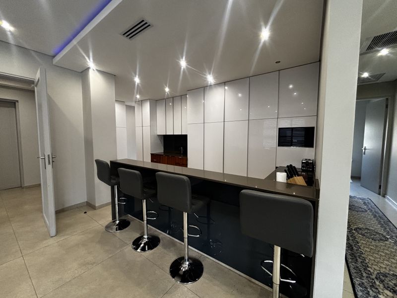 Modern kitchen featuring sleek white cabinetry, a dark stone breakfast bar, recessed ceiling lighting, and large format floor tiles.