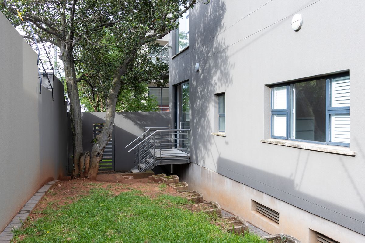 Rear exterior view of the apartment featuring a small garden area, metal staircase access, and modern grey painted facade with aluminium window frames.