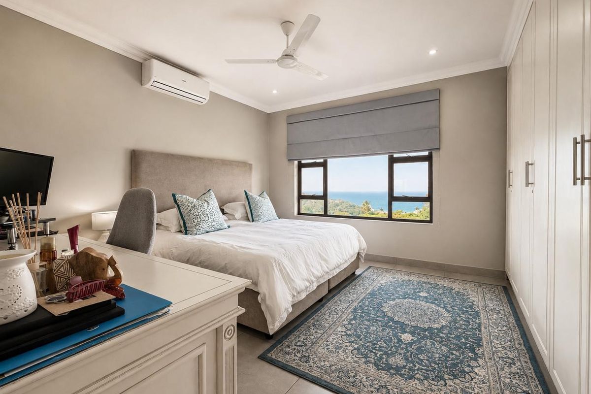 A well-maintained bedroom featuring built-in cupboards, a ceiling fan, air conditioning, and large windows offering scenic ocean views.
