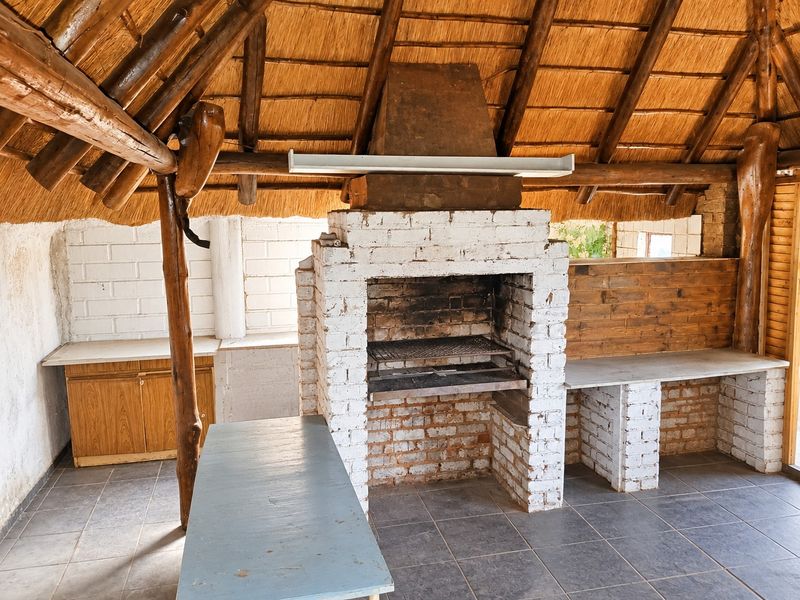 Covered outdoor patio area featuring a traditional thatched roof, built-in brick braai, and tiled flooring, ideal for outdoor entertaining.