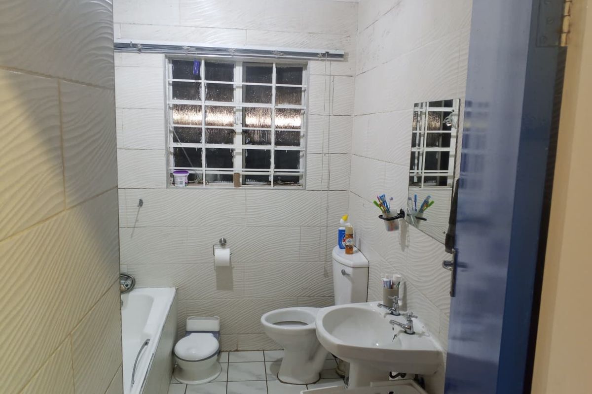 A functional bathroom featuring a bathtub, pedestal basin, toilet, and textured wall tiling with security bars on the window.
