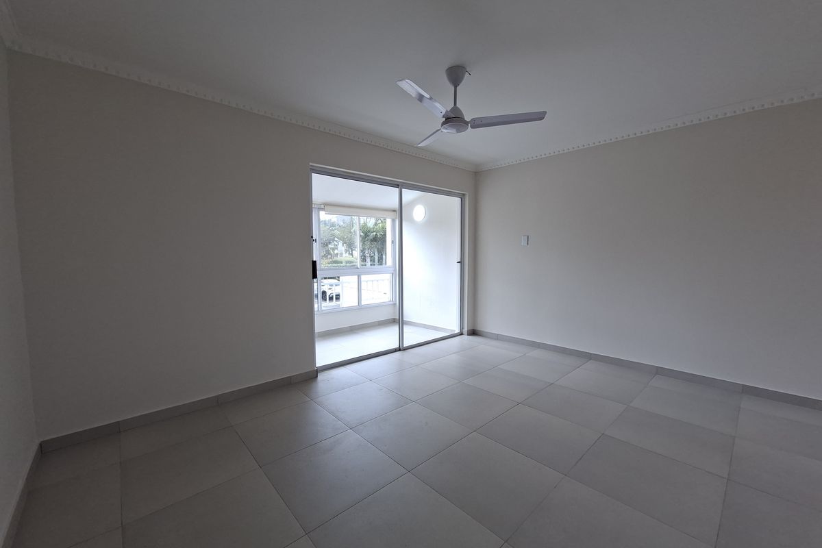 A bright, empty living room featuring neutral tiled flooring, a ceiling fan, and a sliding glass door leading to an outdoor balcony area.