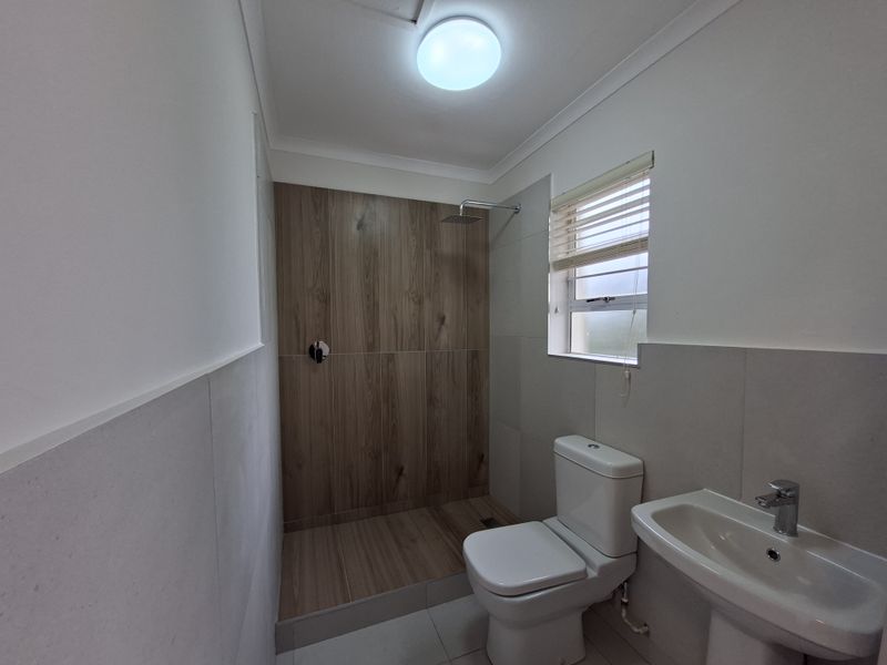A clean, modern bathroom featuring a walk-in shower with wood-look tiling, a pedestal basin, and a toilet, finished with neutral wall tiles.