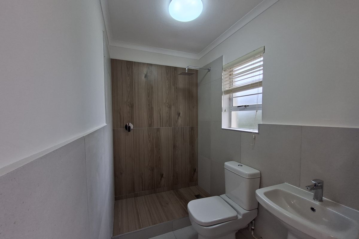 A clean, modern bathroom featuring a walk-in shower with wood-look tiling, a pedestal basin, and a toilet, finished with neutral wall tiles.