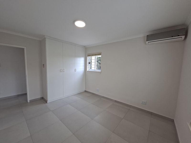 A clean, modern, and empty bedroom featuring neutral tiled flooring, built-in cupboards, and a wall-mounted air conditioning unit.
