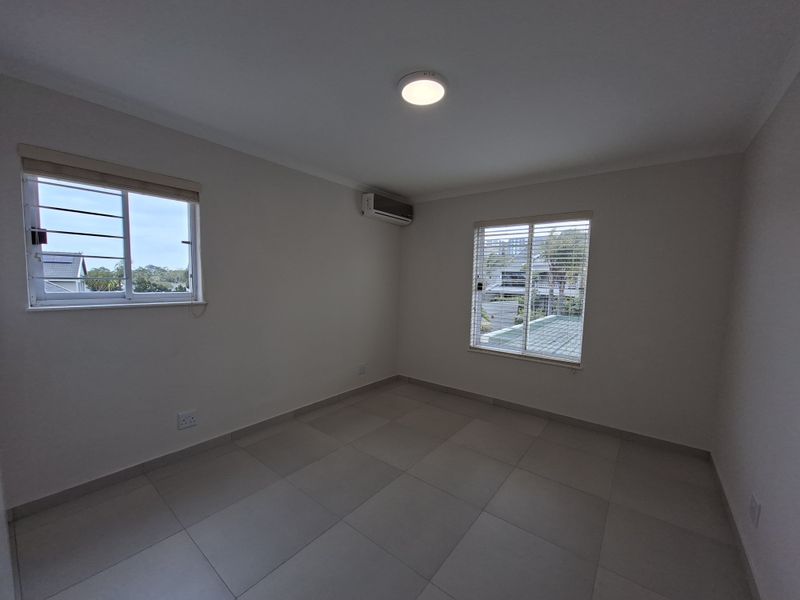 A bright, empty bedroom featuring neutral tiled flooring, an installed air conditioning unit, and windows fitted with security bars and blinds.