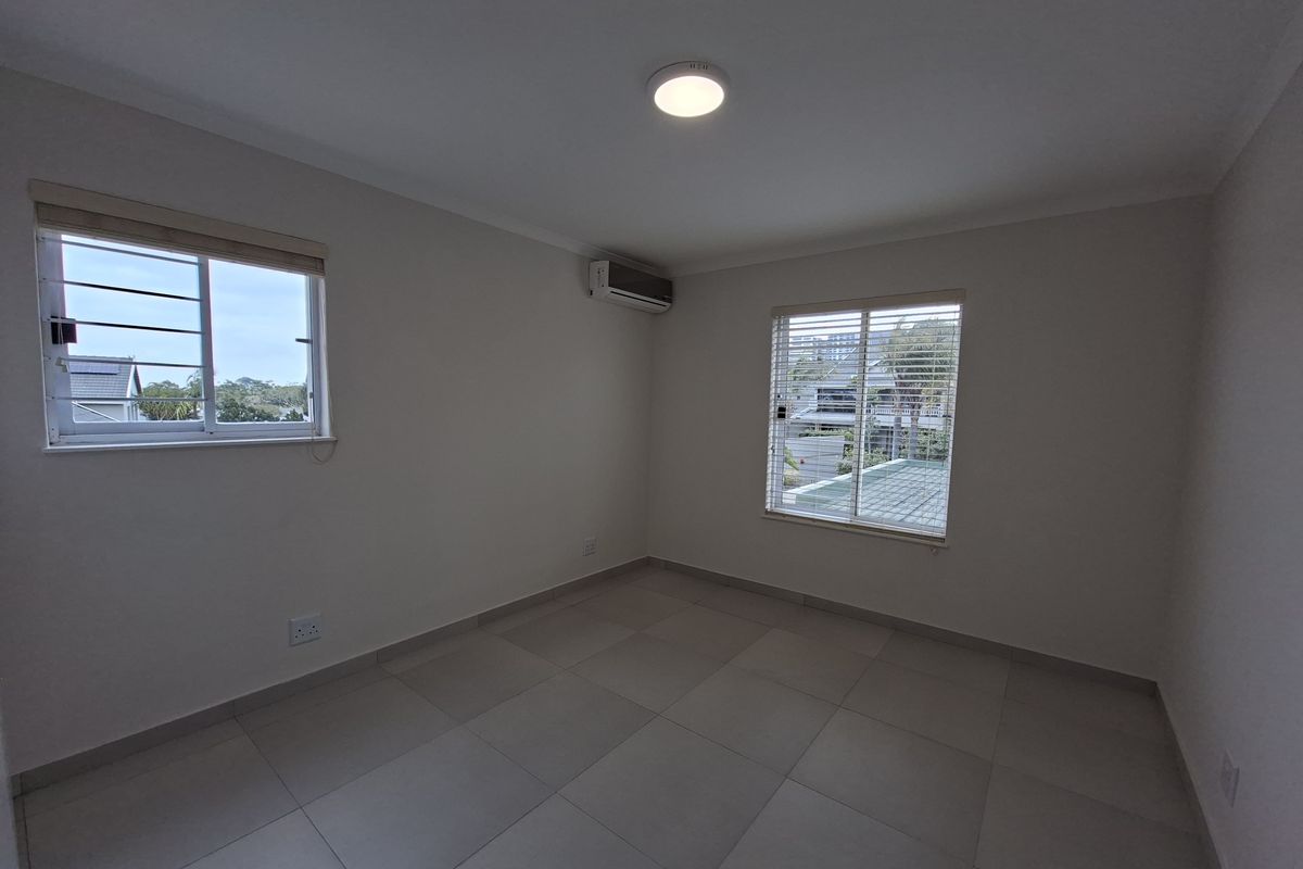 A bright, empty bedroom featuring neutral tiled flooring, an installed air conditioning unit, and windows fitted with security bars and blinds.