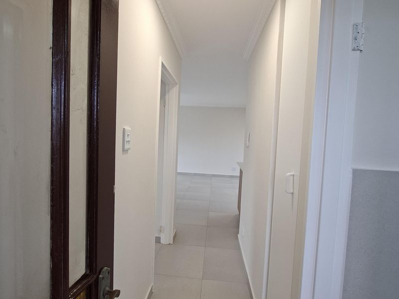 A clean, well-lit hallway featuring neutral tiled flooring, white walls, and classic ceiling cornice detailing, leading into an open-plan living area.