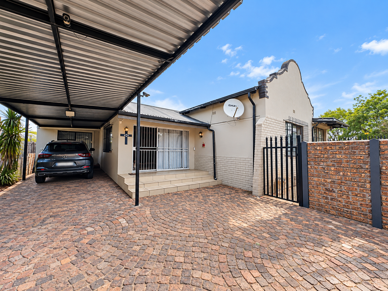 Front exterior view featuring a paved driveway, covered carport, brick boundary wall, and secure entrance with security gates and bars.