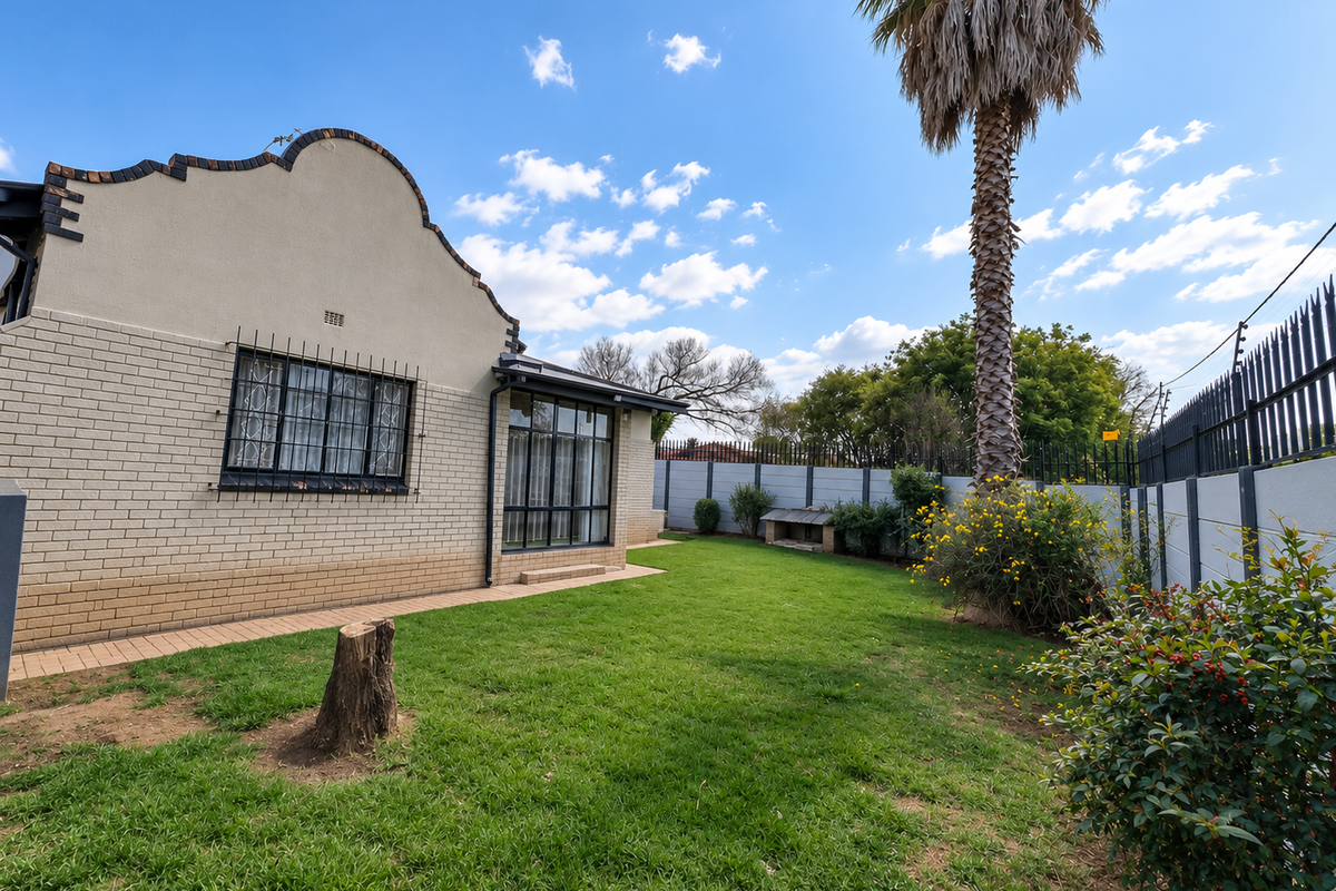 Rear garden view featuring a brick facade house, manicured lawn, established palm tree, and secure perimeter walling.