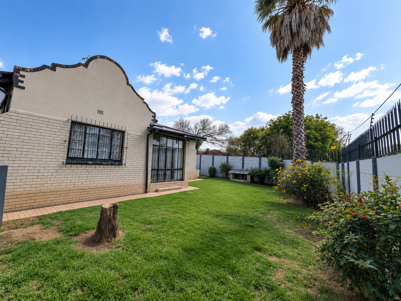 Rear garden view featuring a brick facade house, manicured lawn, established palm tree, and secure perimeter walling.