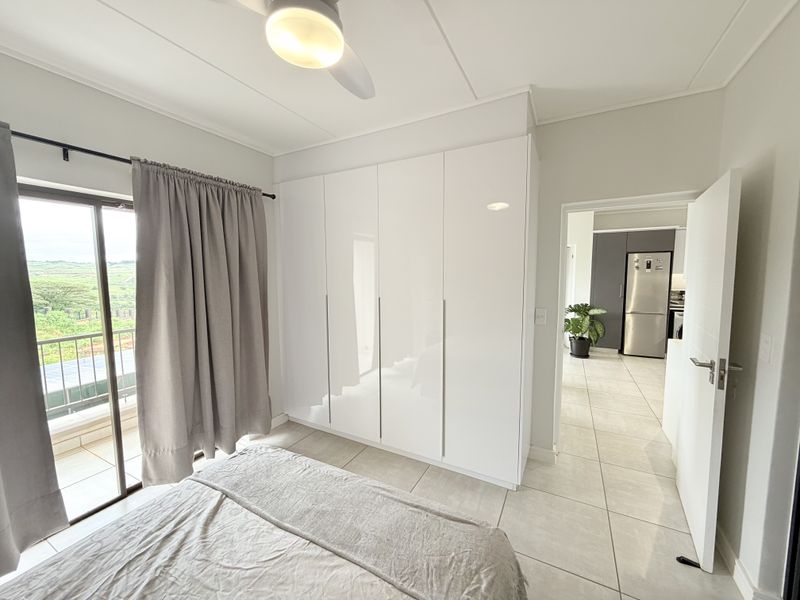 A bright bedroom featuring modern white built-in cupboards, tiled flooring, a ceiling fan, and a sliding glass door leading to a balcony.
