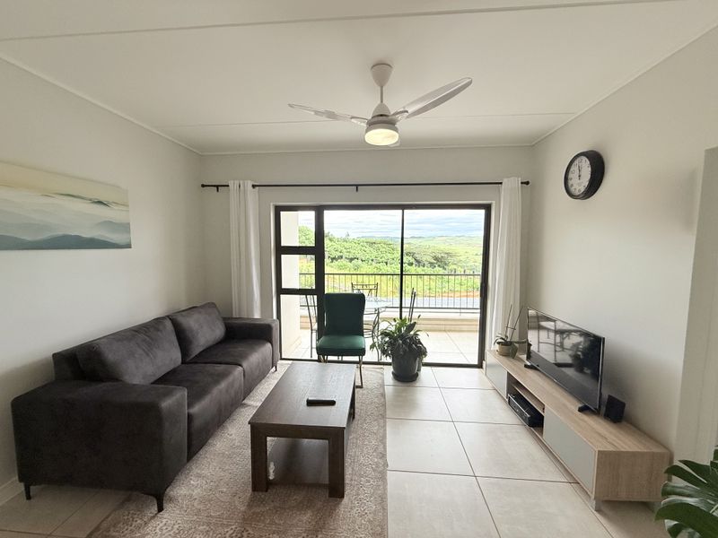 A bright living room featuring tiled flooring, a ceiling fan, and large sliding glass doors that open onto a balcony with scenic views.