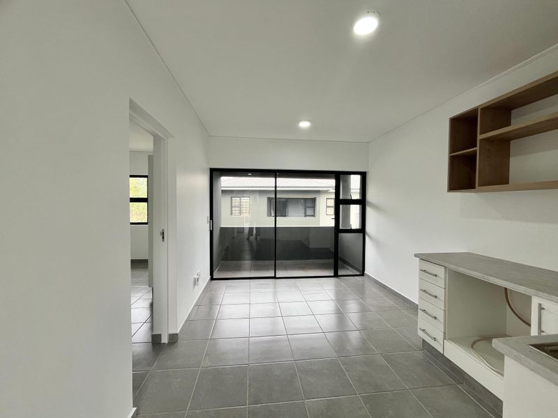 A bright, modern kitchen area featuring neutral tiled flooring, built-in cabinetry with laminate countertops, and a large sliding glass door leading to an outdoor space.