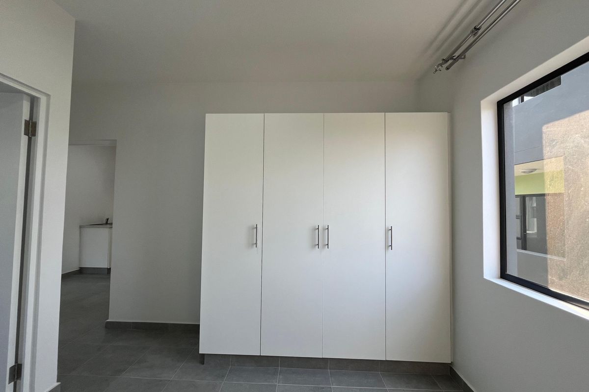 A clean, modern bedroom featuring neutral tiled flooring, a large built-in wardrobe, and a window with a curtain track.