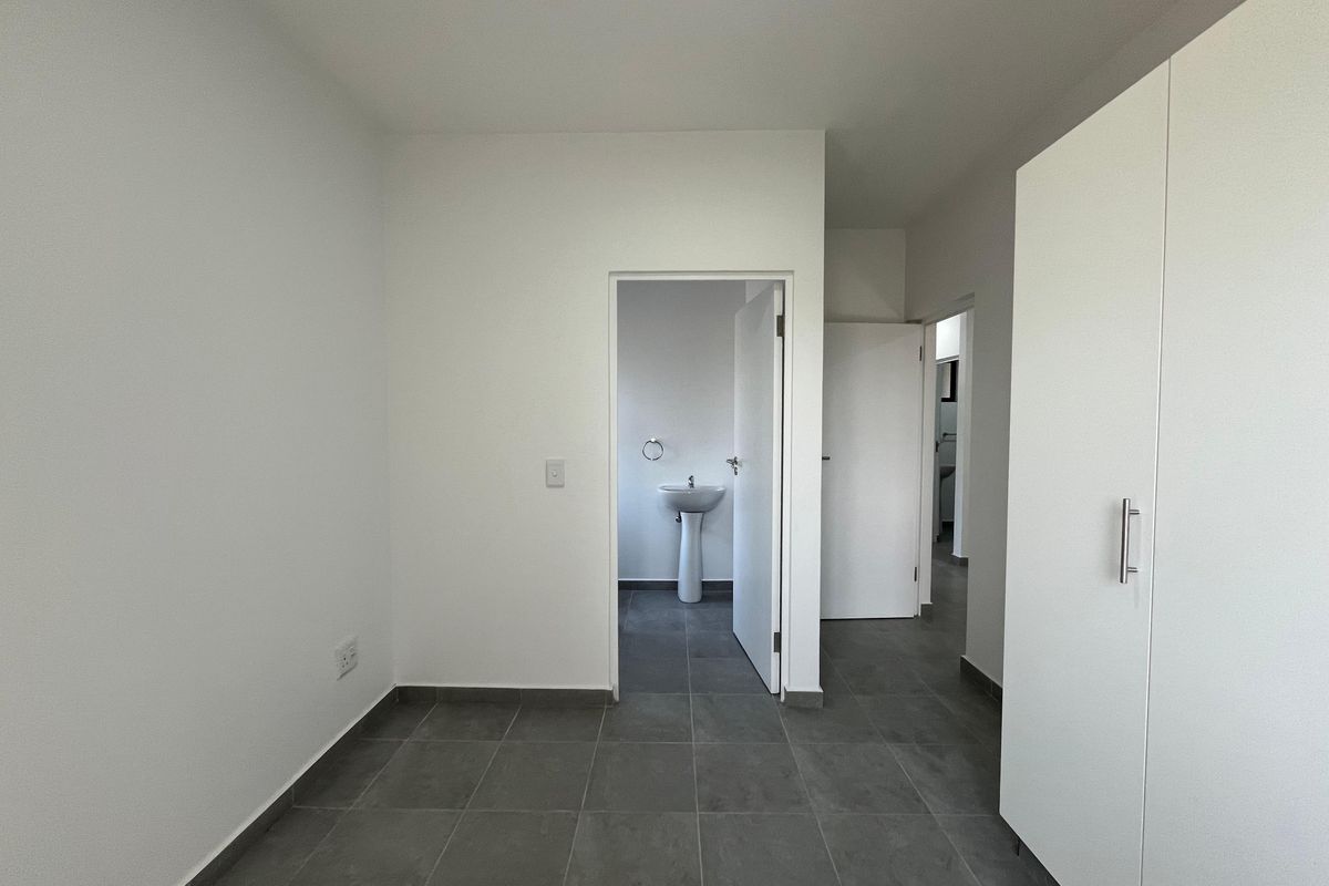 A clean, empty bedroom featuring neutral tiled flooring, built-in wardrobe storage, and direct access to an en-suite bathroom with a pedestal basin.