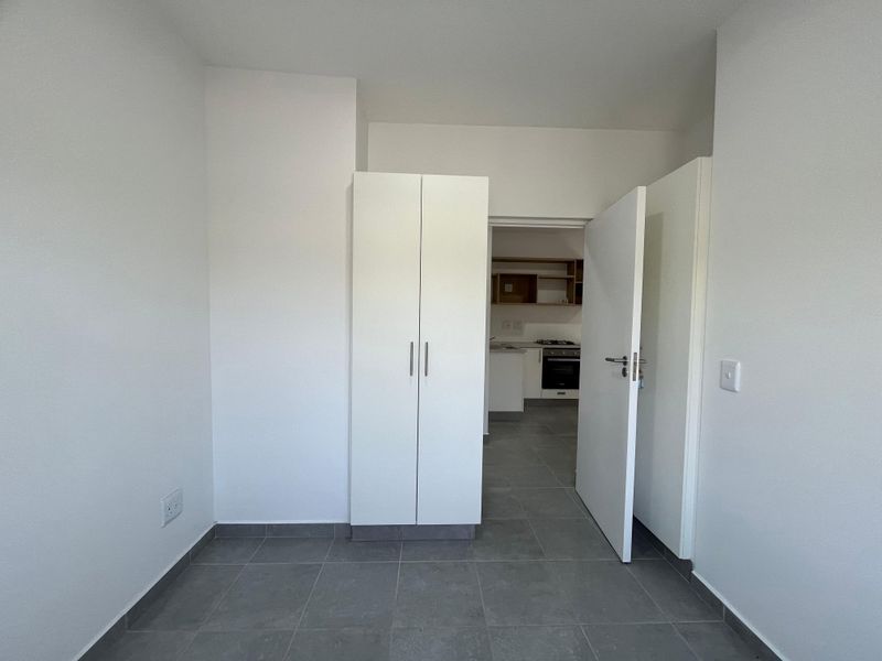 A clean, empty bedroom featuring neutral tiled flooring and a built-in wardrobe, with a view through to the kitchen area.