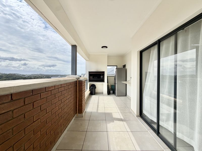 A spacious covered balcony featuring tiled flooring, a built-in braai, and sliding glass doors, offering an ideal outdoor entertainment area with scenic views.