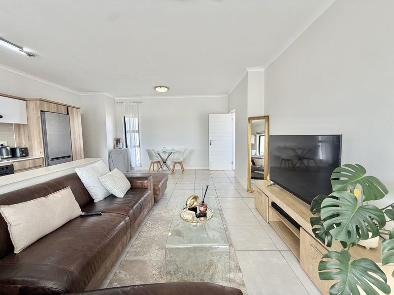 An open-plan living and kitchen area featuring neutral tiled flooring, modern wood-finish cabinetry, and a bright, functional layout suitable for contemporary apartment living.