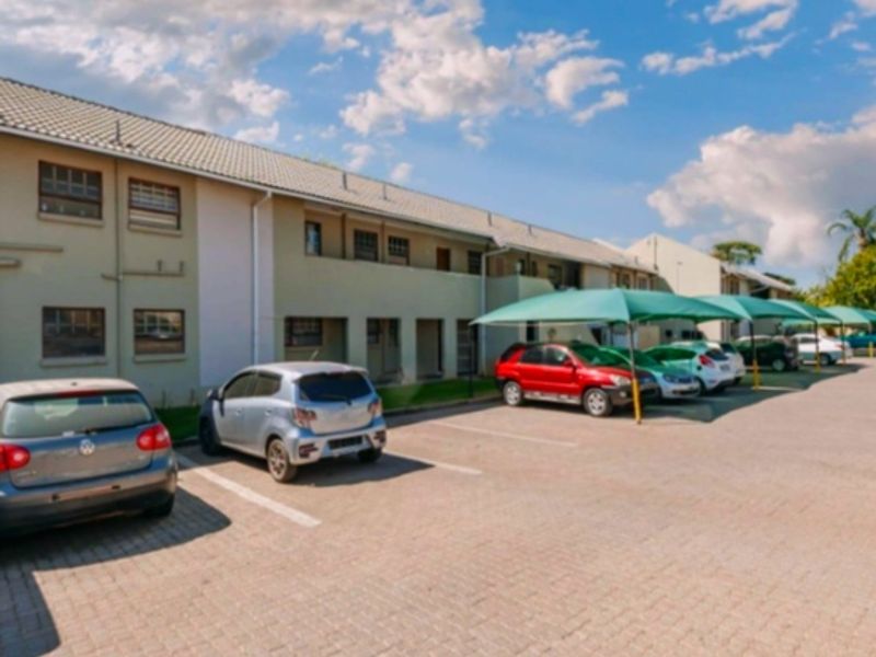 Exterior view of an apartment complex featuring a paved parking area with shade cloth carports and a multi-level building facade.