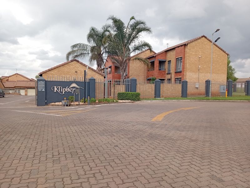 Street view of the Klipberg townhouse complex entrance, featuring brick facade buildings, paved driveway, and secure perimeter fencing.