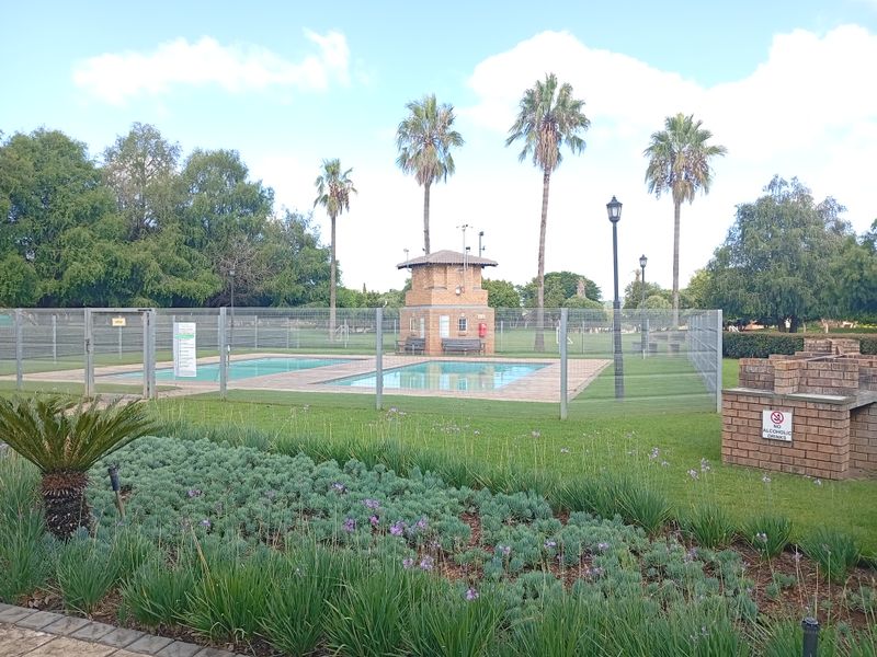 Communal pool area featuring a brick pool house, perimeter fencing, and a built-in brick braai set within a well-maintained landscaped garden.