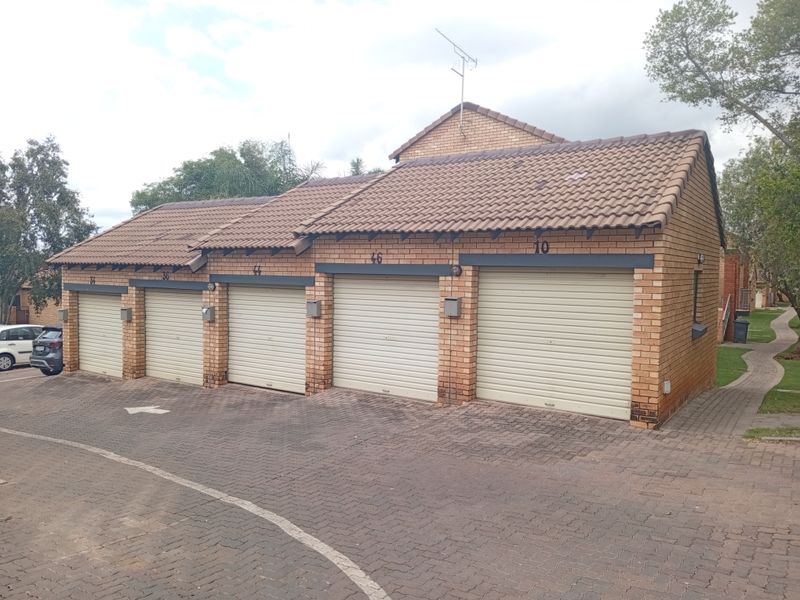 A row of brick-built garages with tiled roofing and roller doors, situated in a paved residential complex setting.