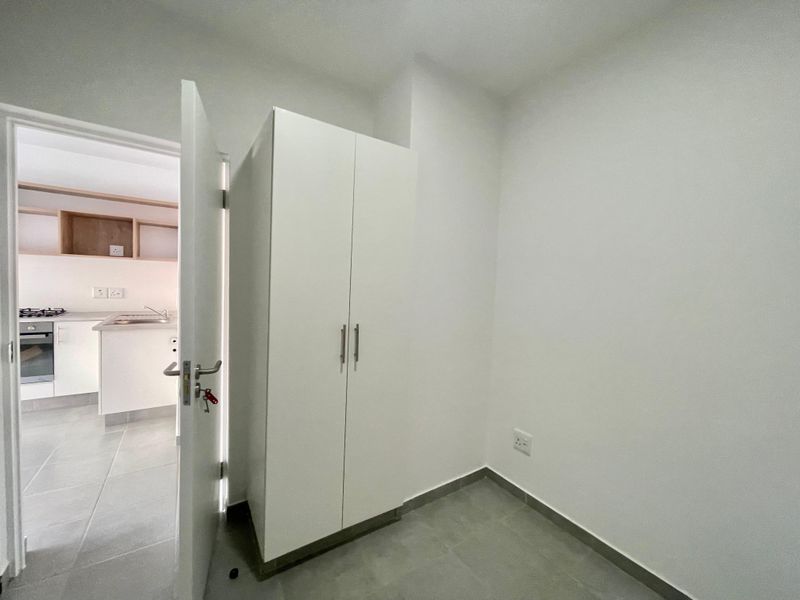 A clean, empty bedroom featuring neutral tiled flooring and a modern white built-in cupboard, with a view into the adjacent kitchen area.