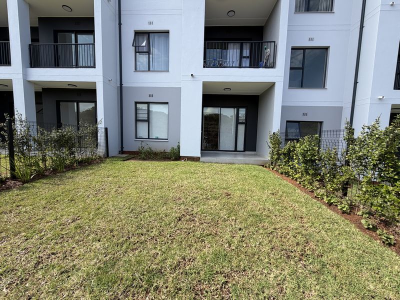 Rear view of a modern apartment building featuring a private ground-floor garden, tiled patio area, and upper-level balconies with metal railings.