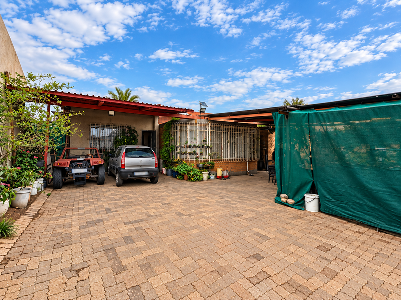 A paved rear courtyard area featuring a covered patio structure, security bars on windows, and brick walling.