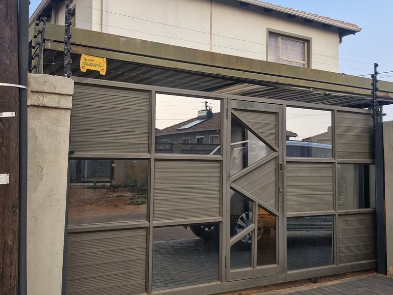 Front entrance view featuring a modern sliding gate, perimeter wall with electric fencing, and an integrated carport structure.