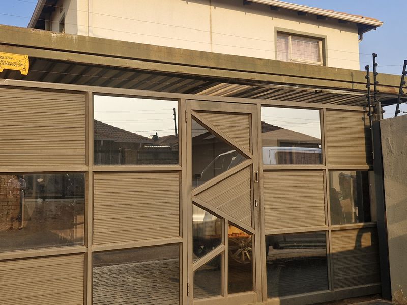 Front entrance view featuring a modern metal gate, perimeter wall with electric fencing, and a covered carport area.