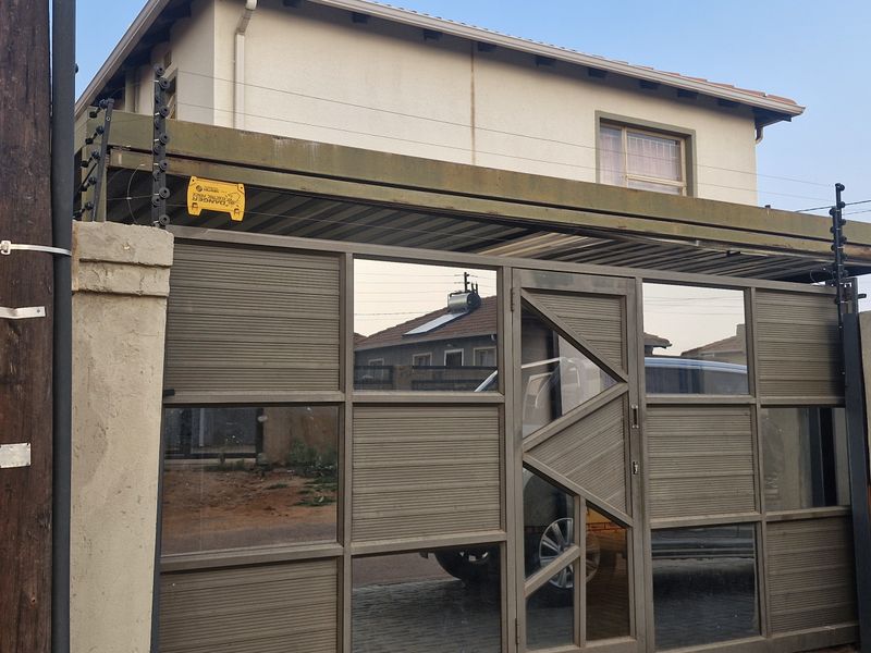 Front view of the property featuring a modern metal sliding gate, integrated carport, and perimeter masonry wall with installed electric security fencing.