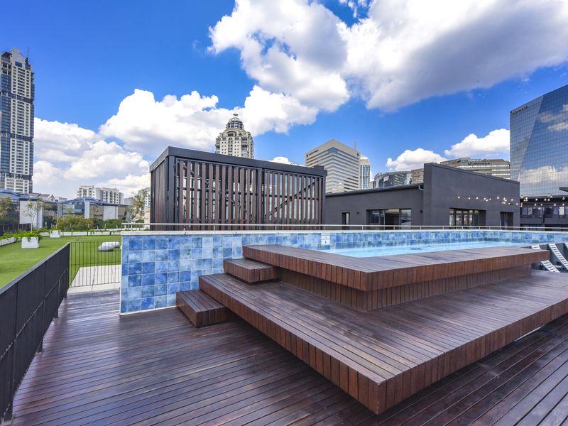 Rooftop pool area featuring raised timber decking, blue tiled surround, and artificial lawn with expansive city skyline views.