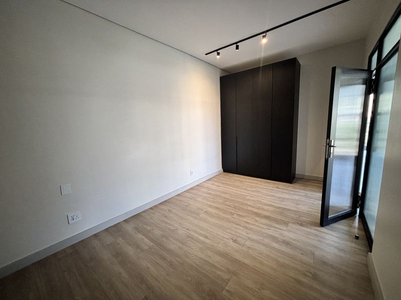 A modern, empty bedroom featuring light wood-look laminate flooring, a sleek black built-in wardrobe, and contemporary track lighting.