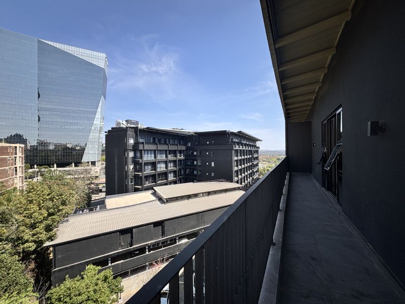 A modern, covered balcony featuring dark tiled flooring and a metal balustrade, offering an elevated urban view of the surrounding neighborhood.
